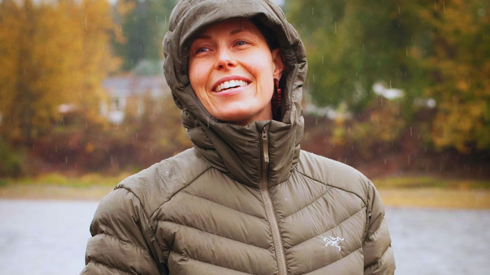 A woman in a green jacket smiles while standing in the rain, showcasing her joy despite the weather.