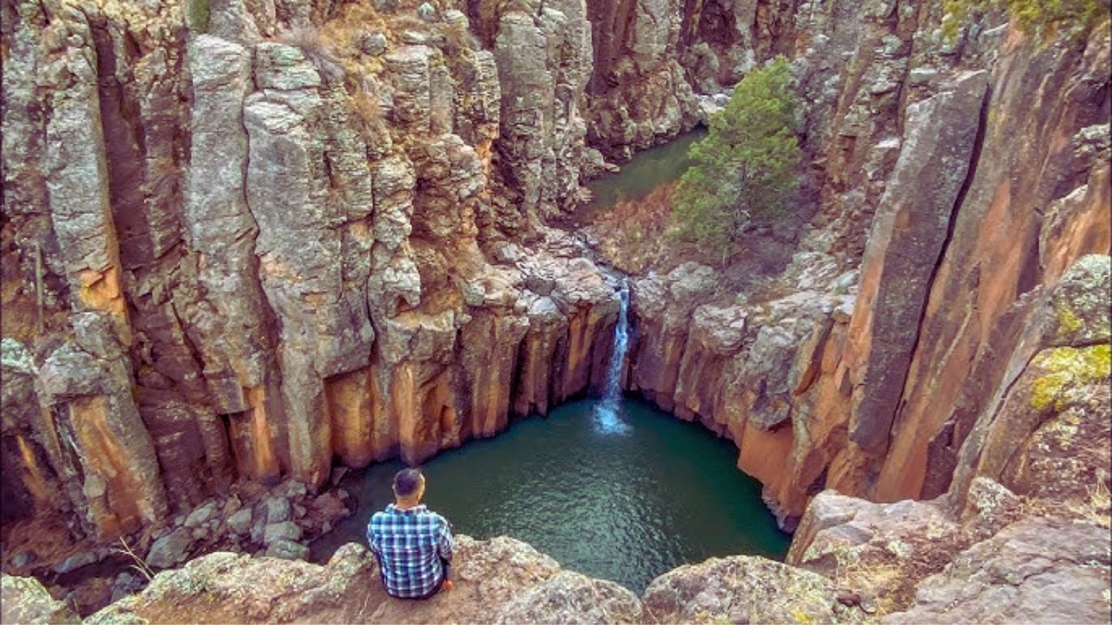 A man sits on a cliff's edge, gazing at a cascading waterfall below him.