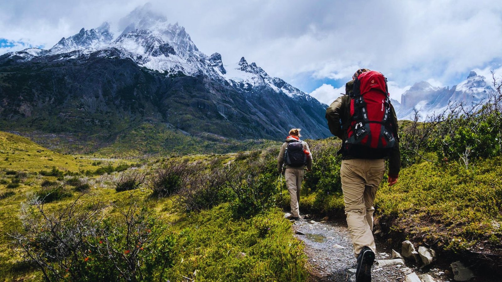Two hikers with backpacks walking along a mountain trail surrounded by lush greenery and rocky terrain