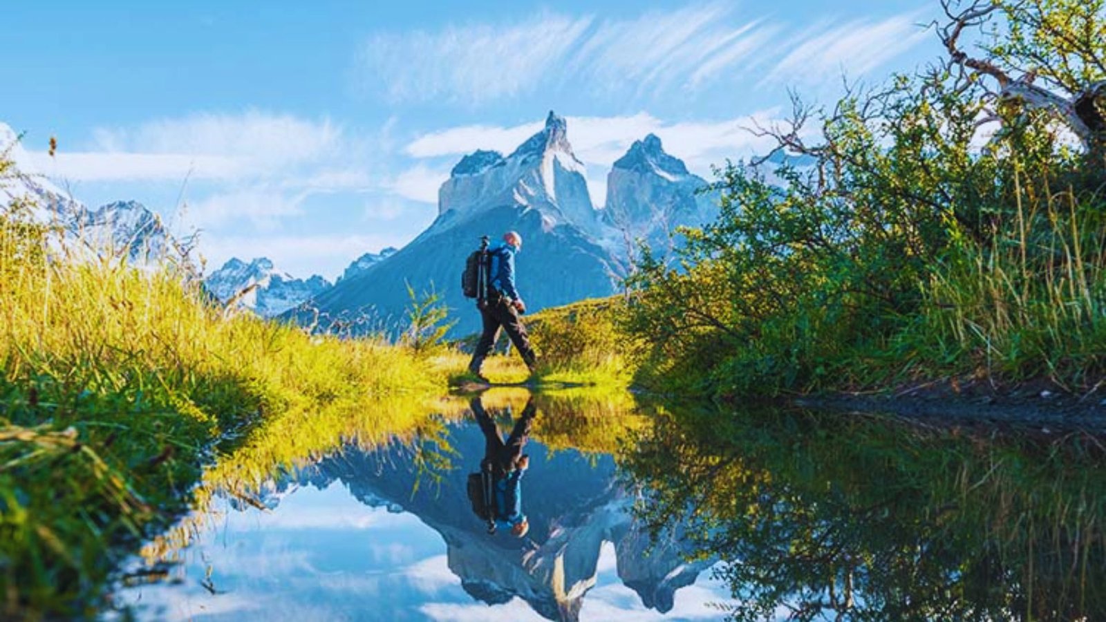 A man walks along a scenic path, with serene water reflecting the surrounding mountains in the background.
