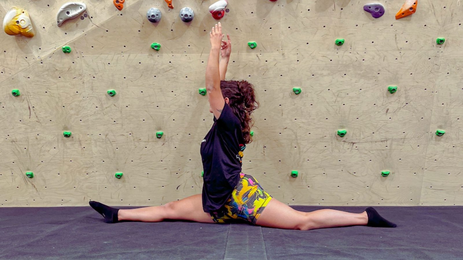 A woman performs a split on a climbing wall, showcasing her flexibility and strength in a dynamic pose.