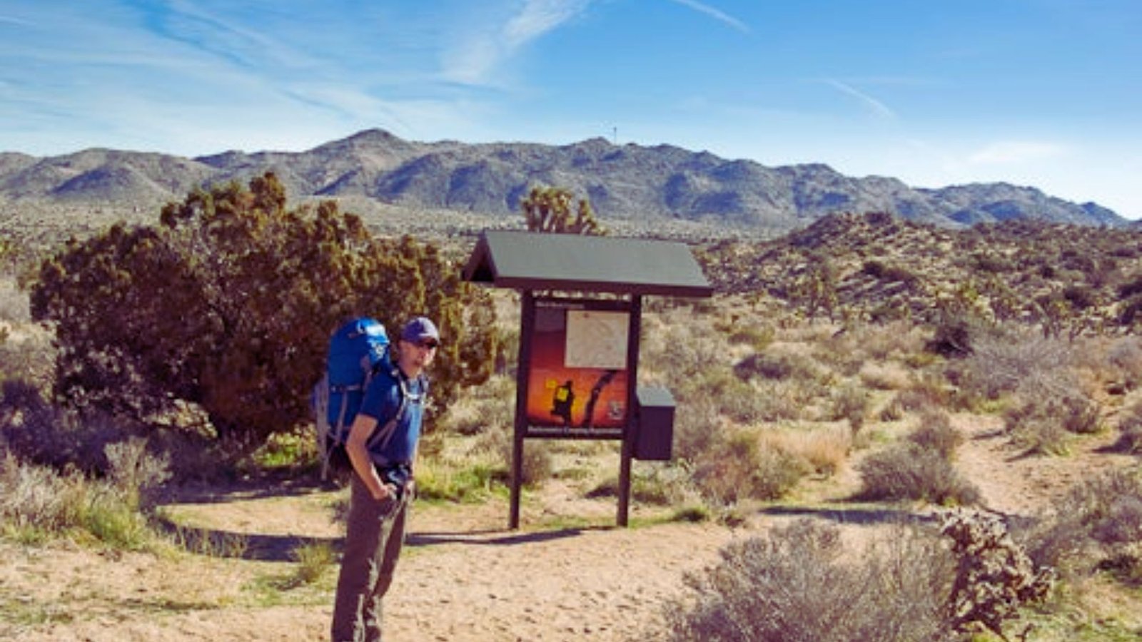 A man with a backpack stands at the entrance of Joshua Tree National Monument, surrounded by desert landscape.