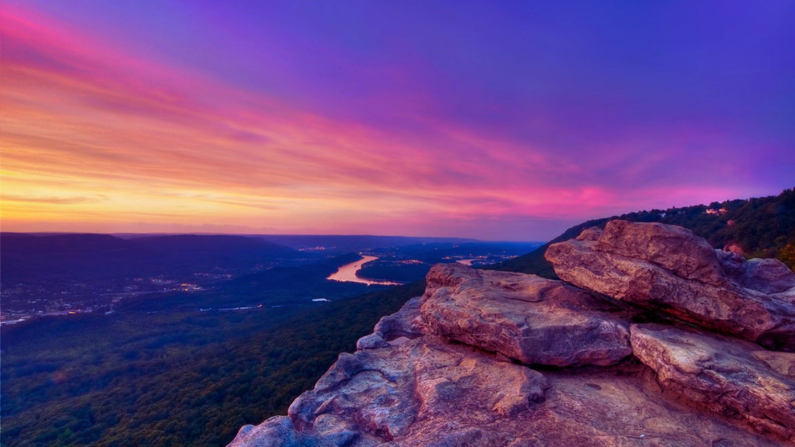 A stunning sunset paints the sky with colors above the mountains and river, creating a serene landscape.