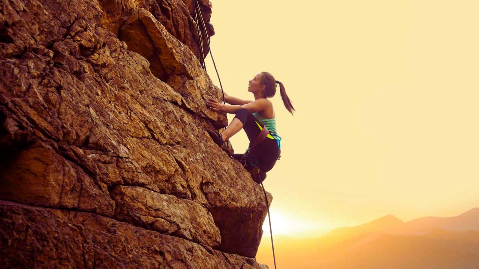 A woman climbs a mountain at sunset, silhouetted against a vibrant orange and purple sky.