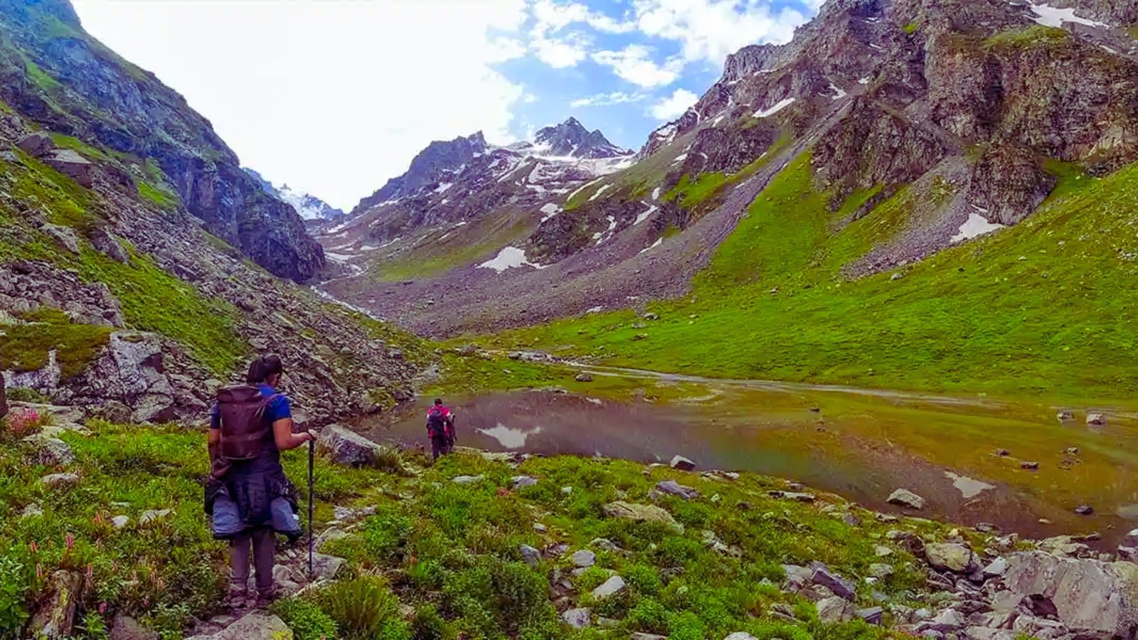  Two hikers ascend a mountain trail, with a serene lake visible in the background under a clear blue sky.