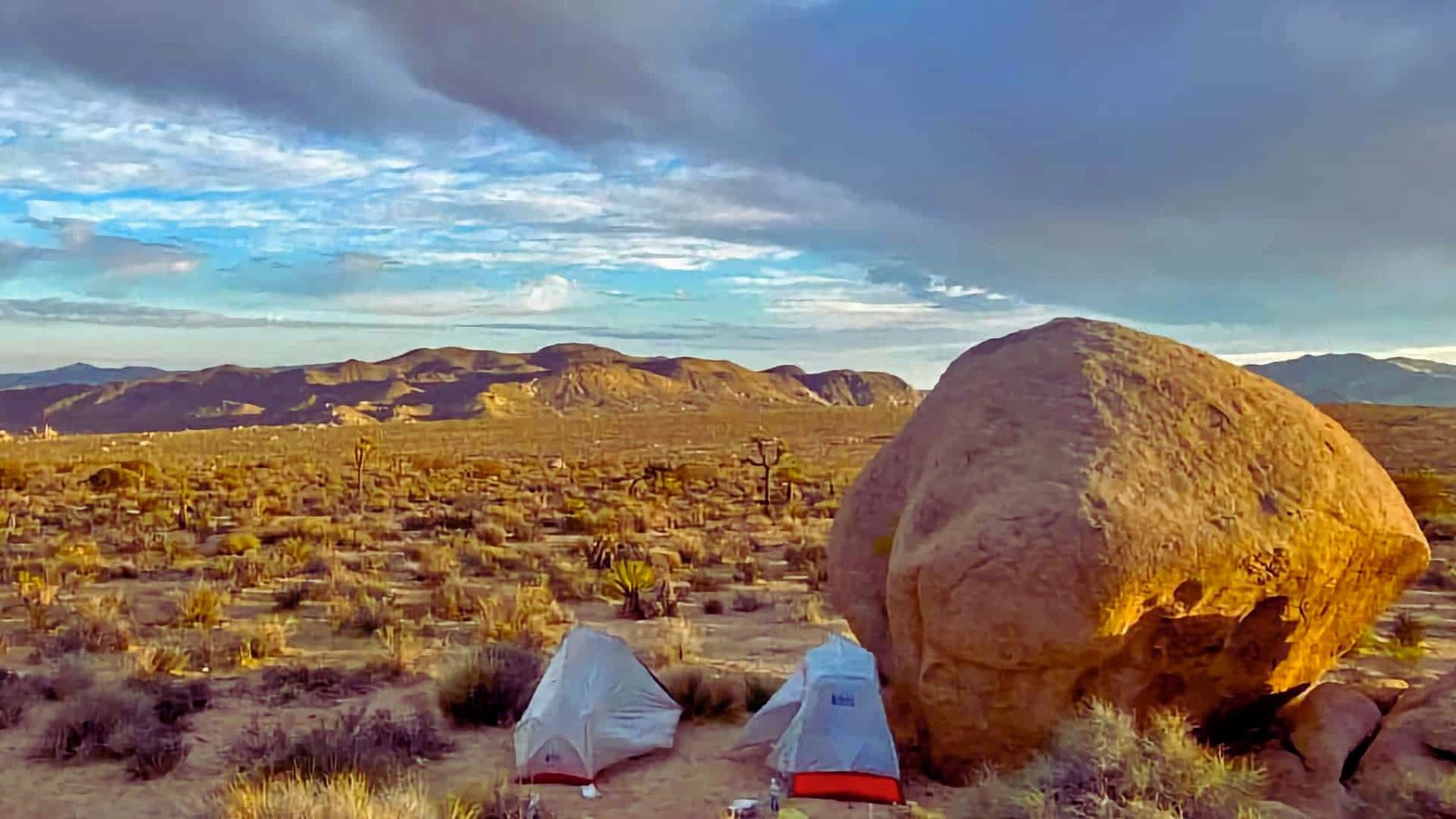 A tent is pitched in the desert, accompanied by a large rock formation nearby under a clear blue sky.