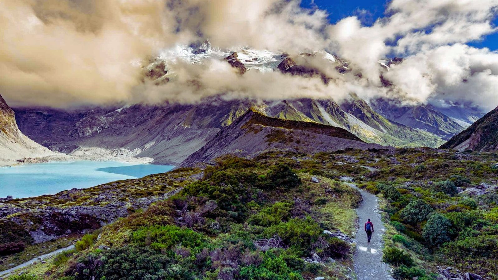 A man walks along a scenic path beside a lake, with mountains rising majestically in the background.