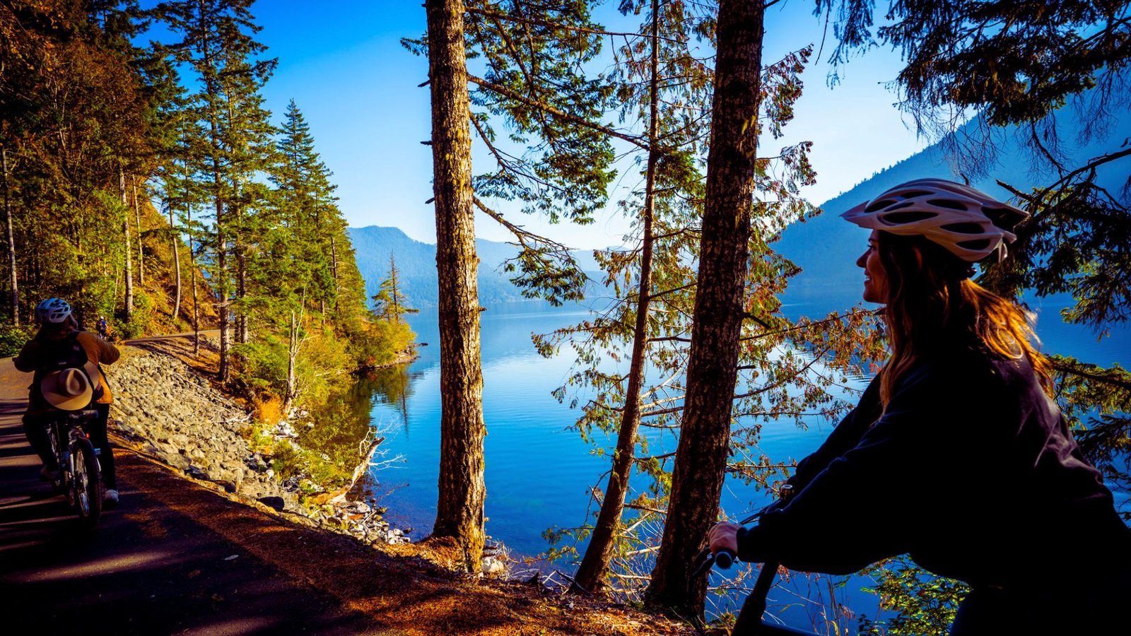 Two cyclists riding along a lakeside trail, enjoying the serene view of the water and lush surroundings.