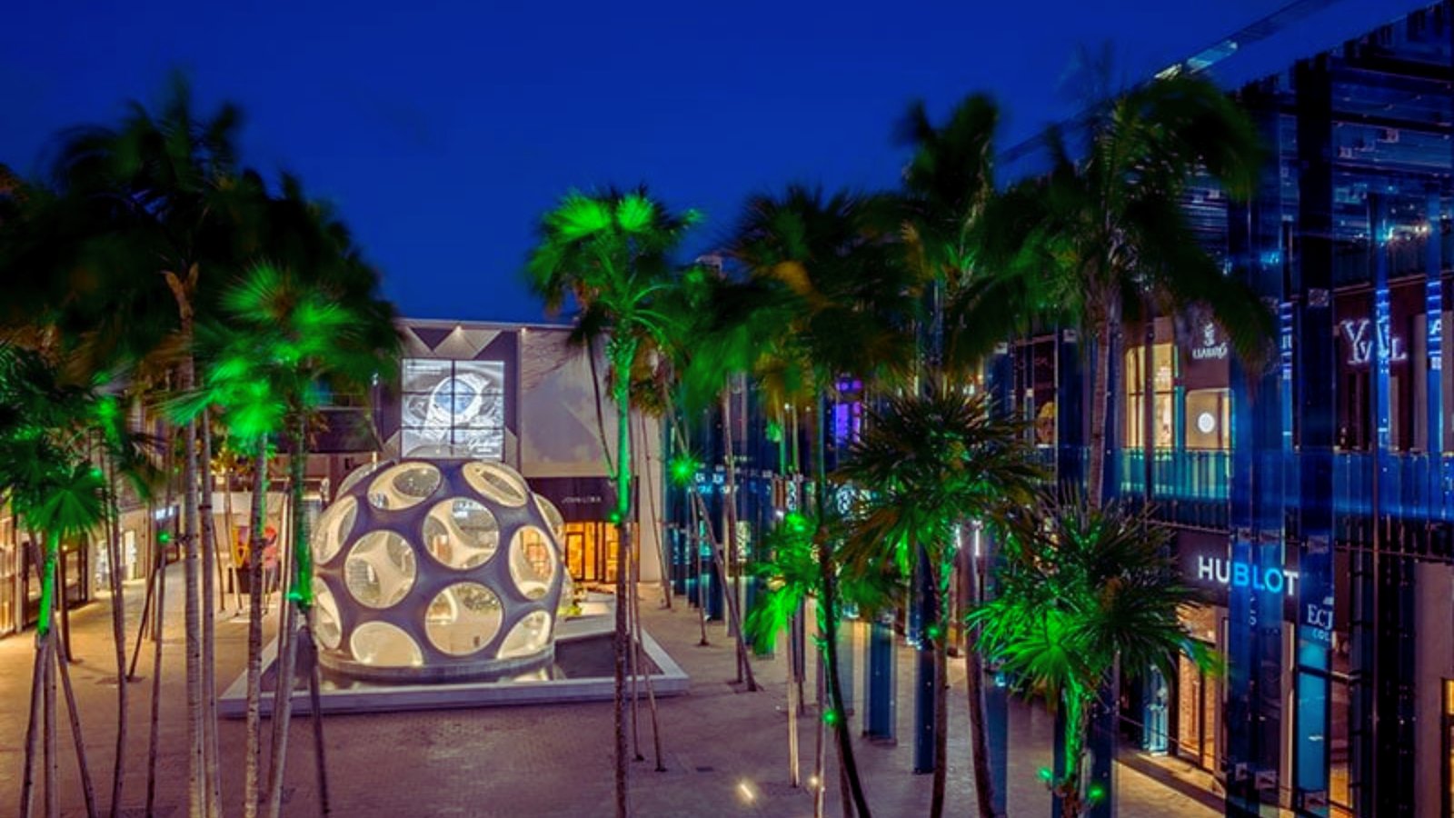 A shopping mall exterior illuminated at night, showcasing bright lights and a bustling atmosphere.