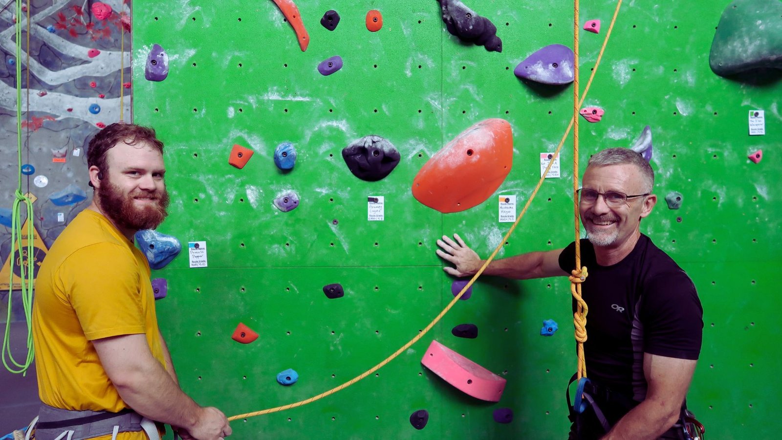 Two men stand together at a climbing wall, discussing their next moves.