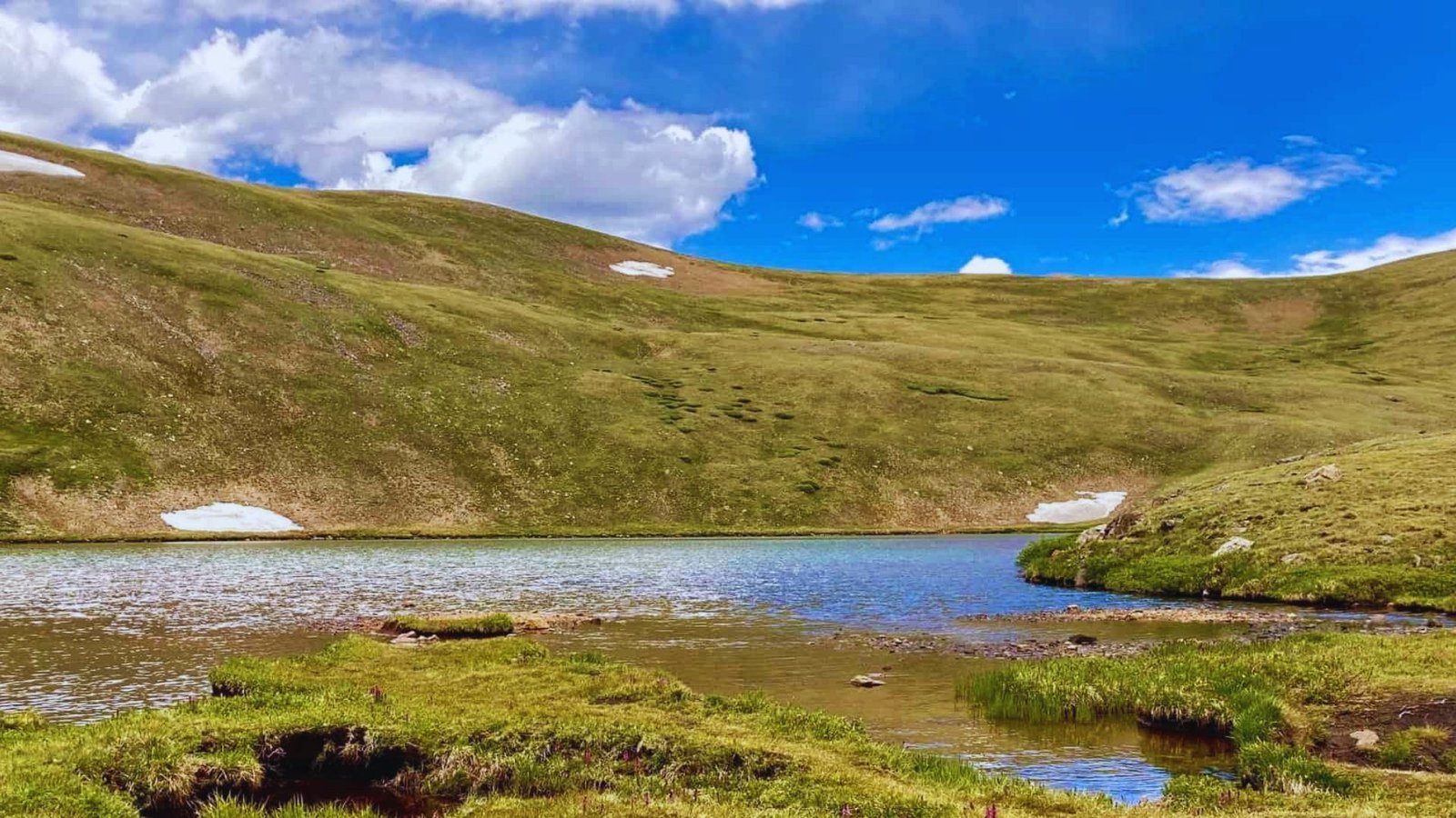 A serene lake nestled among grassy hills, with majestic snow-capped mountains in the background.