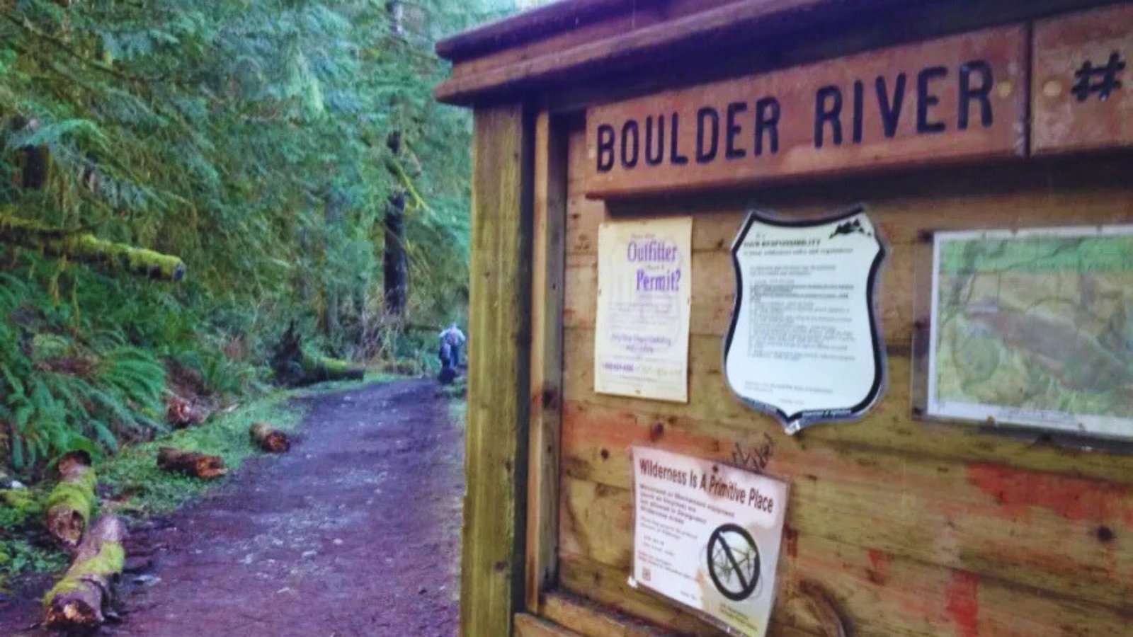  A sign reading "Boulder River" stands in a lush forest, indicating the trail parking information nearby.

