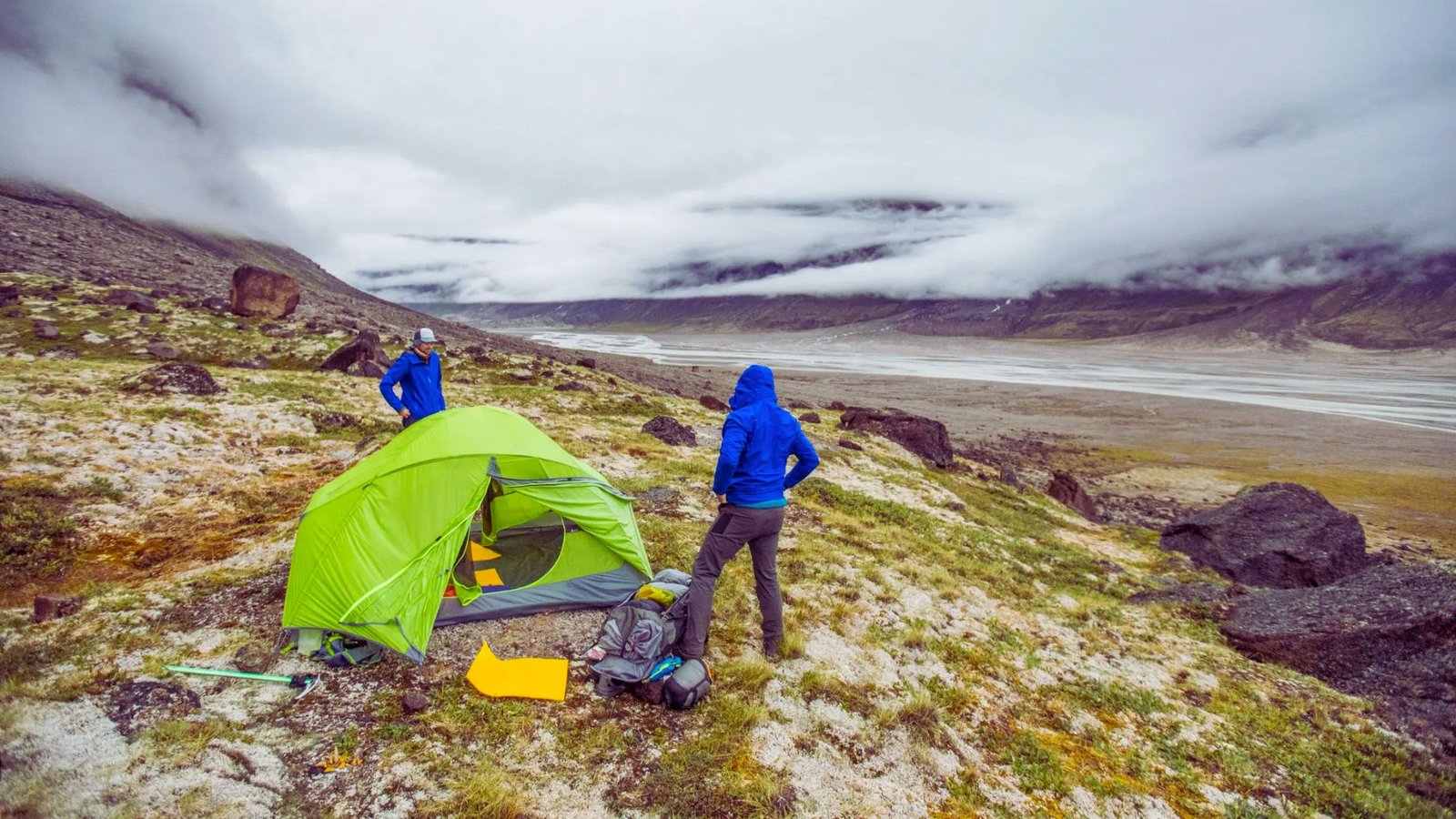 Two people stand beside a tent, surrounded by majestic mountains under a clear blue sky.