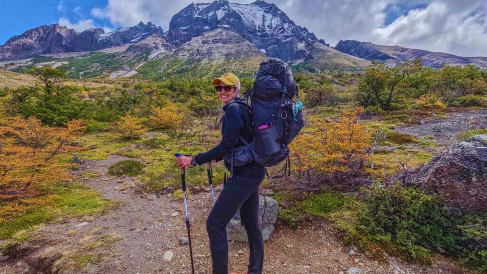 A woman with a backpack and hiking poles stands in the mountains, surrounded by scenic peaks and clear blue skies.