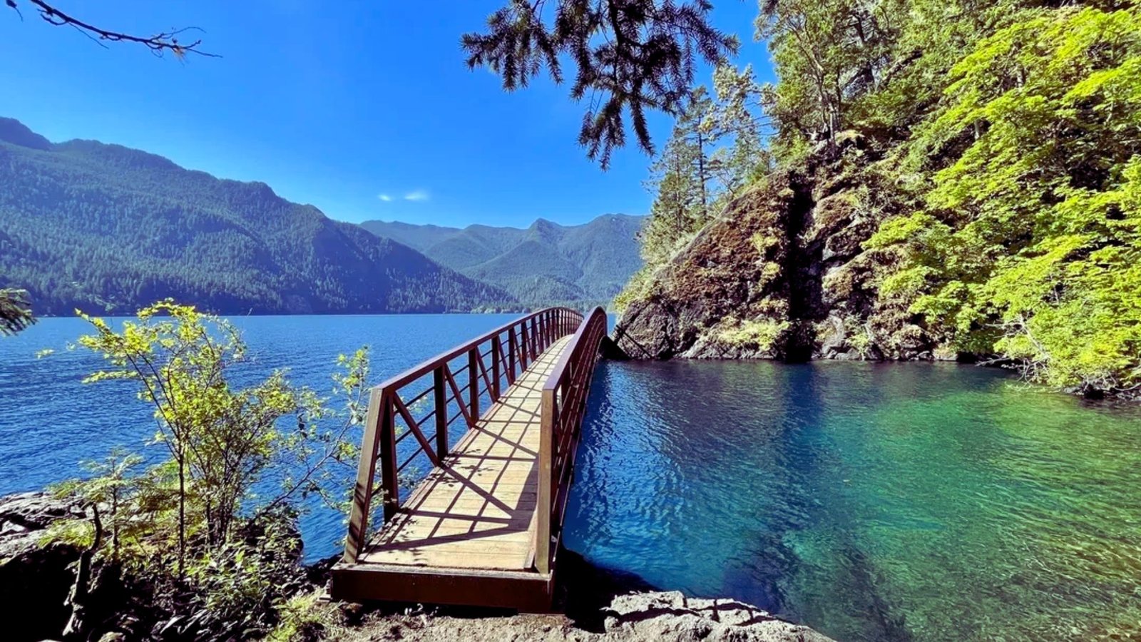 A wooden bridge spans a serene lake, framed by majestic mountains in the background.