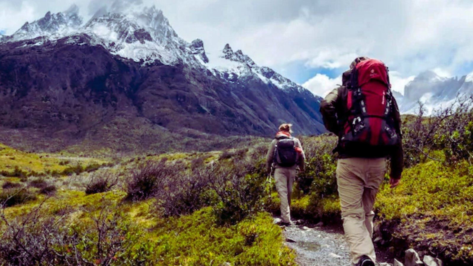  Two hikers with backpacks walking through a scenic mountain range under a clear blue sky.