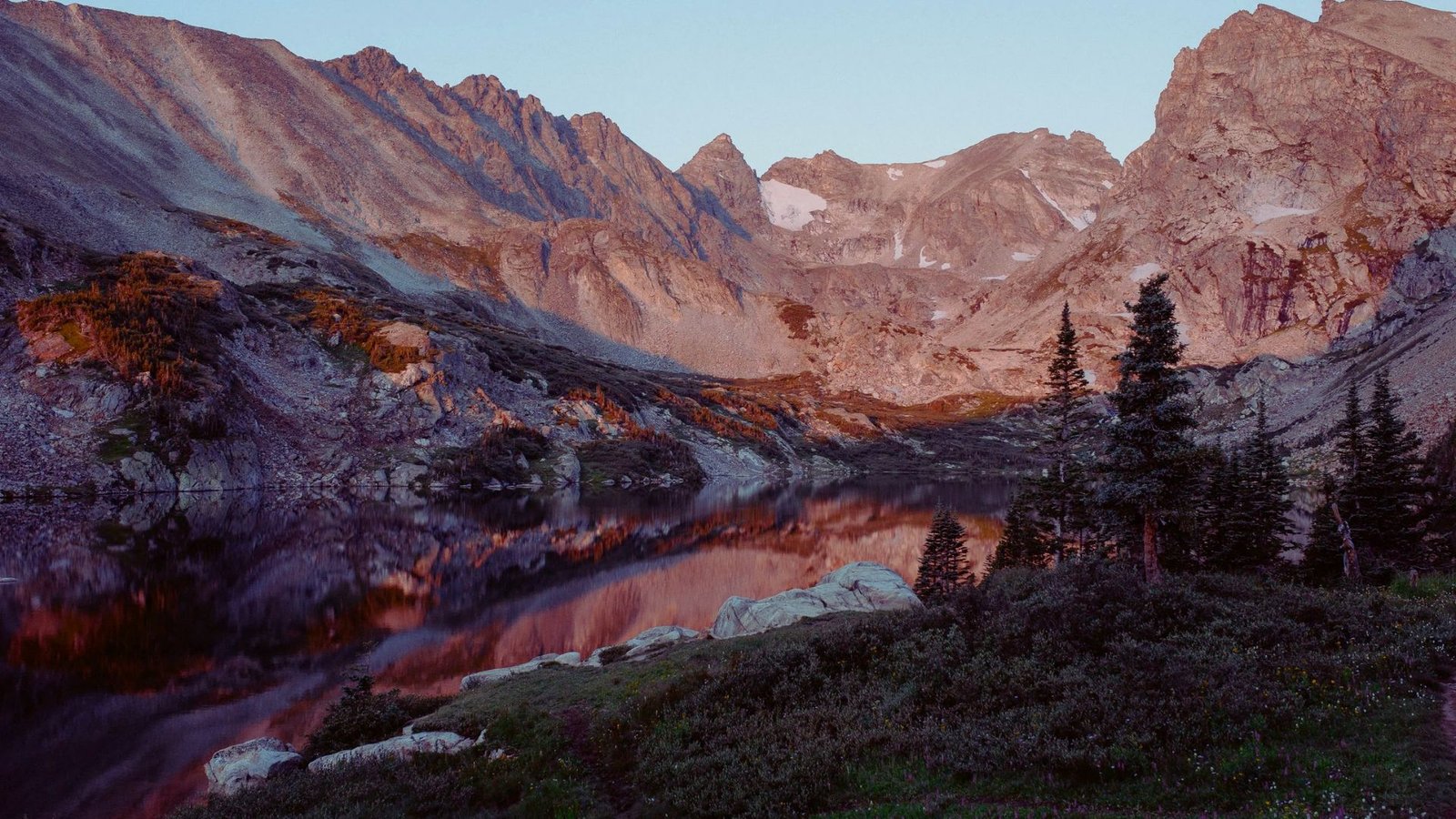 A serene mountain lake nestled among lush trees and towering mountains under a clear blue sky.