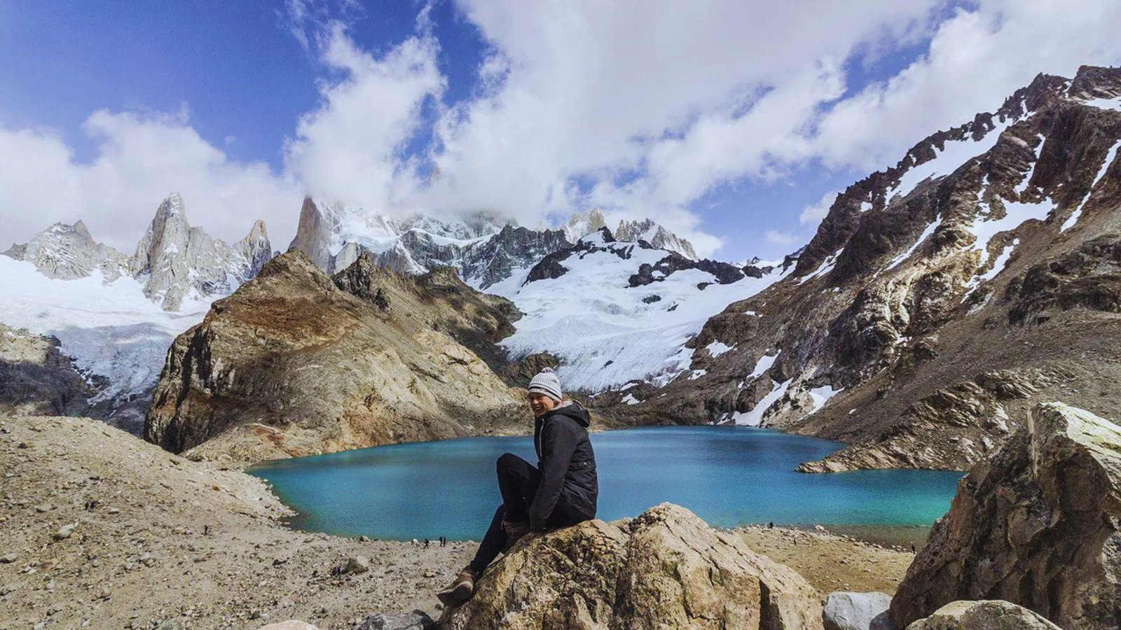 A man sits on a rock by a serene lake, surrounded by nature and reflecting on the calm waters.