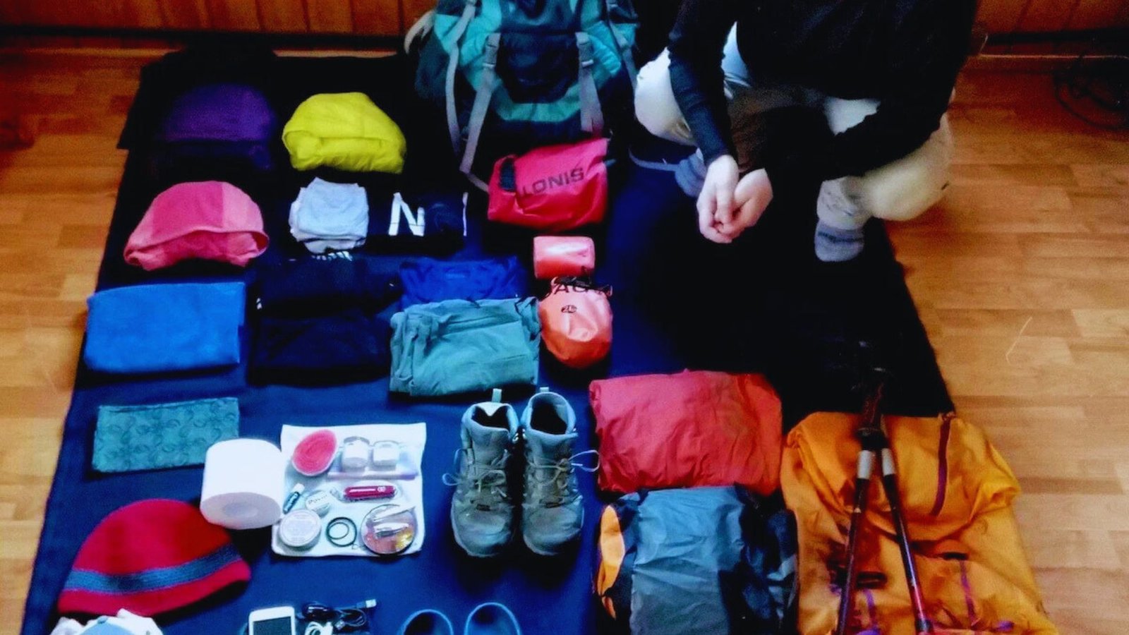 A person sitting on the floor surrounded by a backpack and various items, appearing relaxed and engaged in their surroundings.
