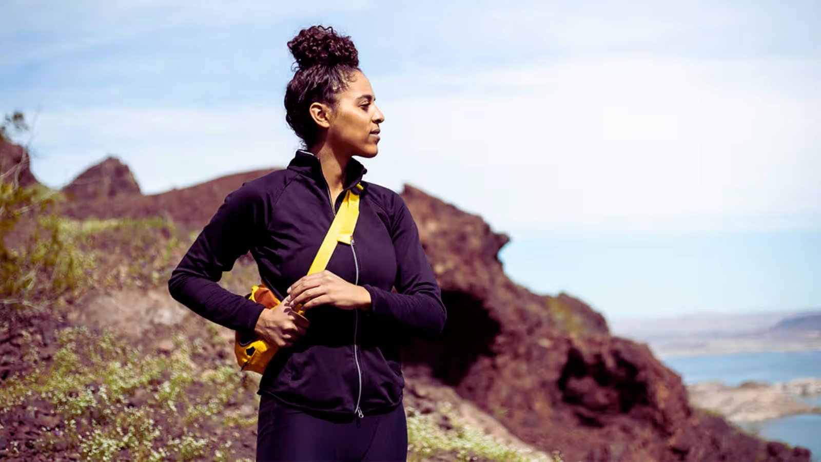 A woman stands on a mountain peak, wearing a yellow backpack, with a scenic view of the landscape behind her.
