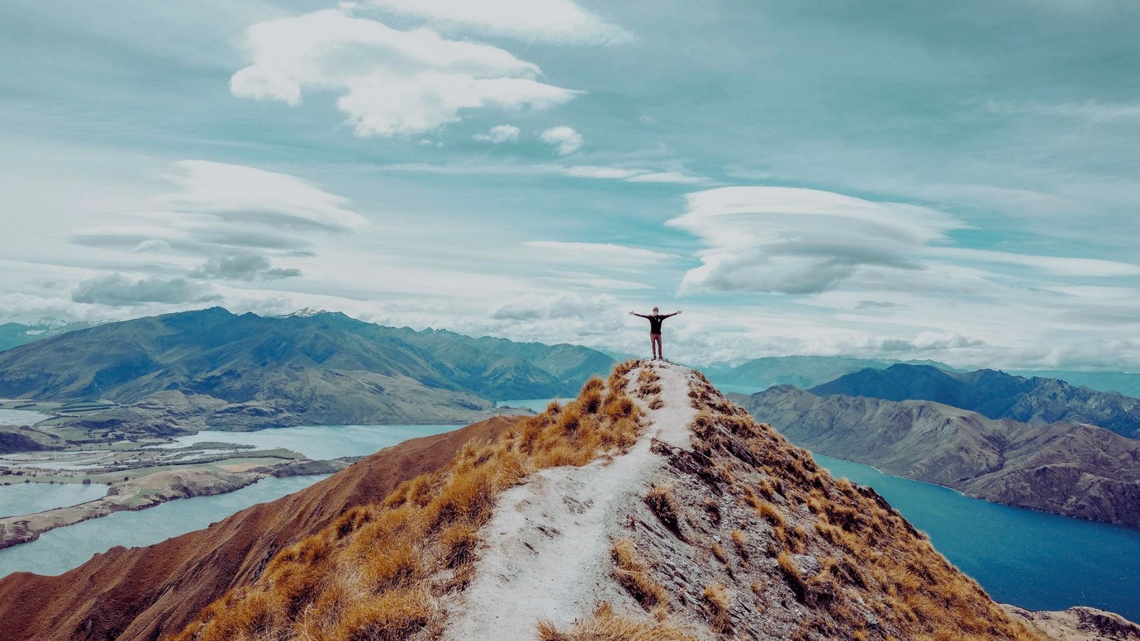 Man standing triumphantly on a mountain peak with arms raised, celebrating a successful climb against a clear blue sky.