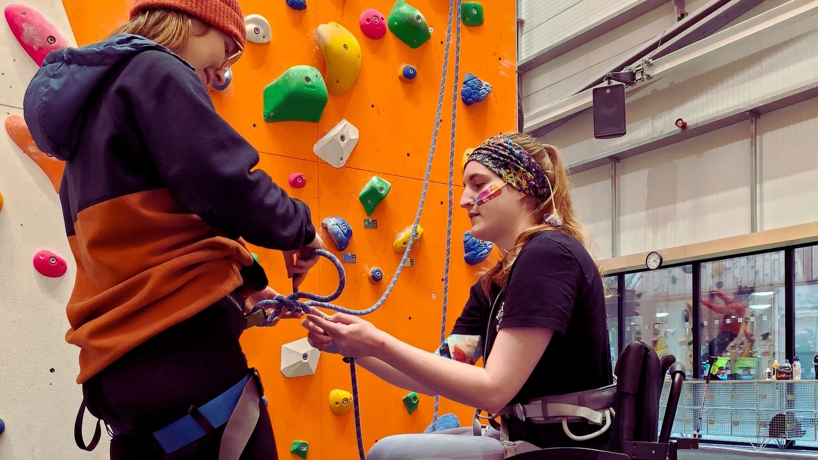 woman in a wheelchair holds a rope while another woman climbs a wall, showcasing teamwork and determination.