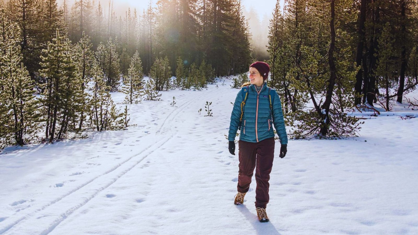 A woman walks through a snowy forest, surrounded by tall trees blanketed in white snow.