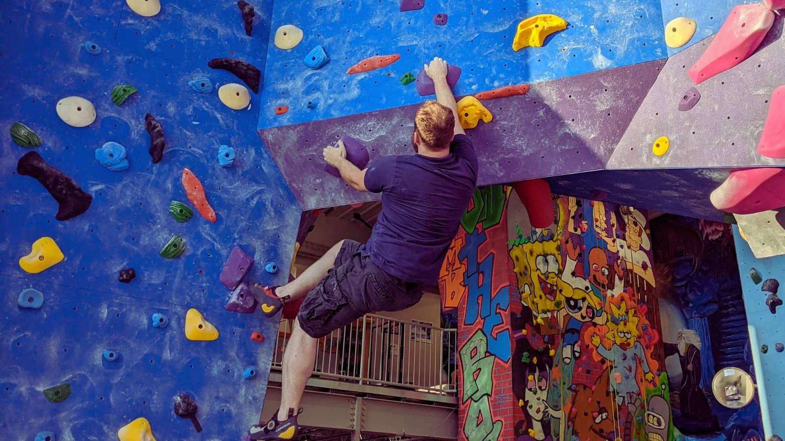 A man climbing a rock wall inside a gym, focused on reaching the next hold.