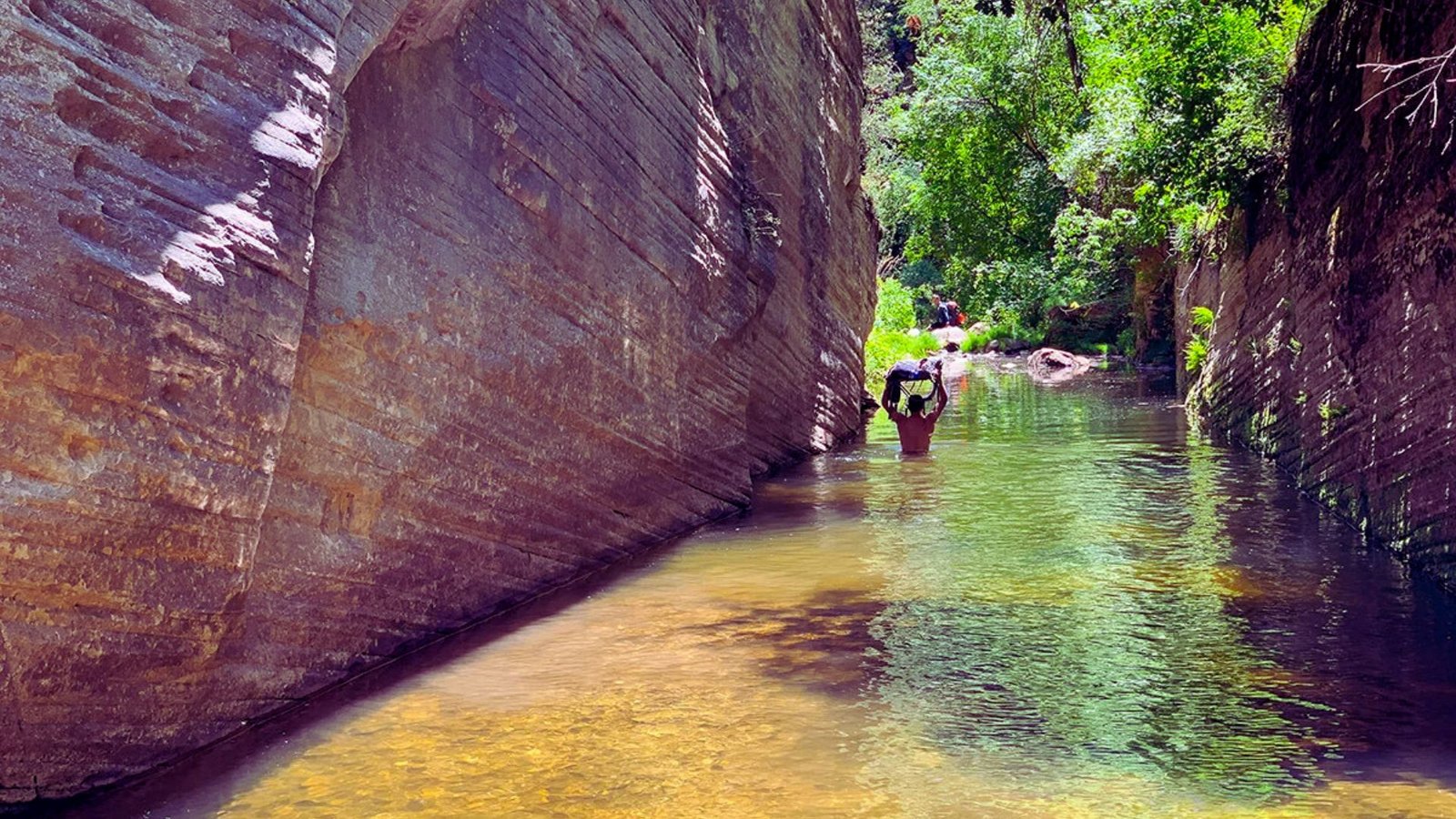 A man walks through a narrow canyon, surrounded by towering rock walls on either side.