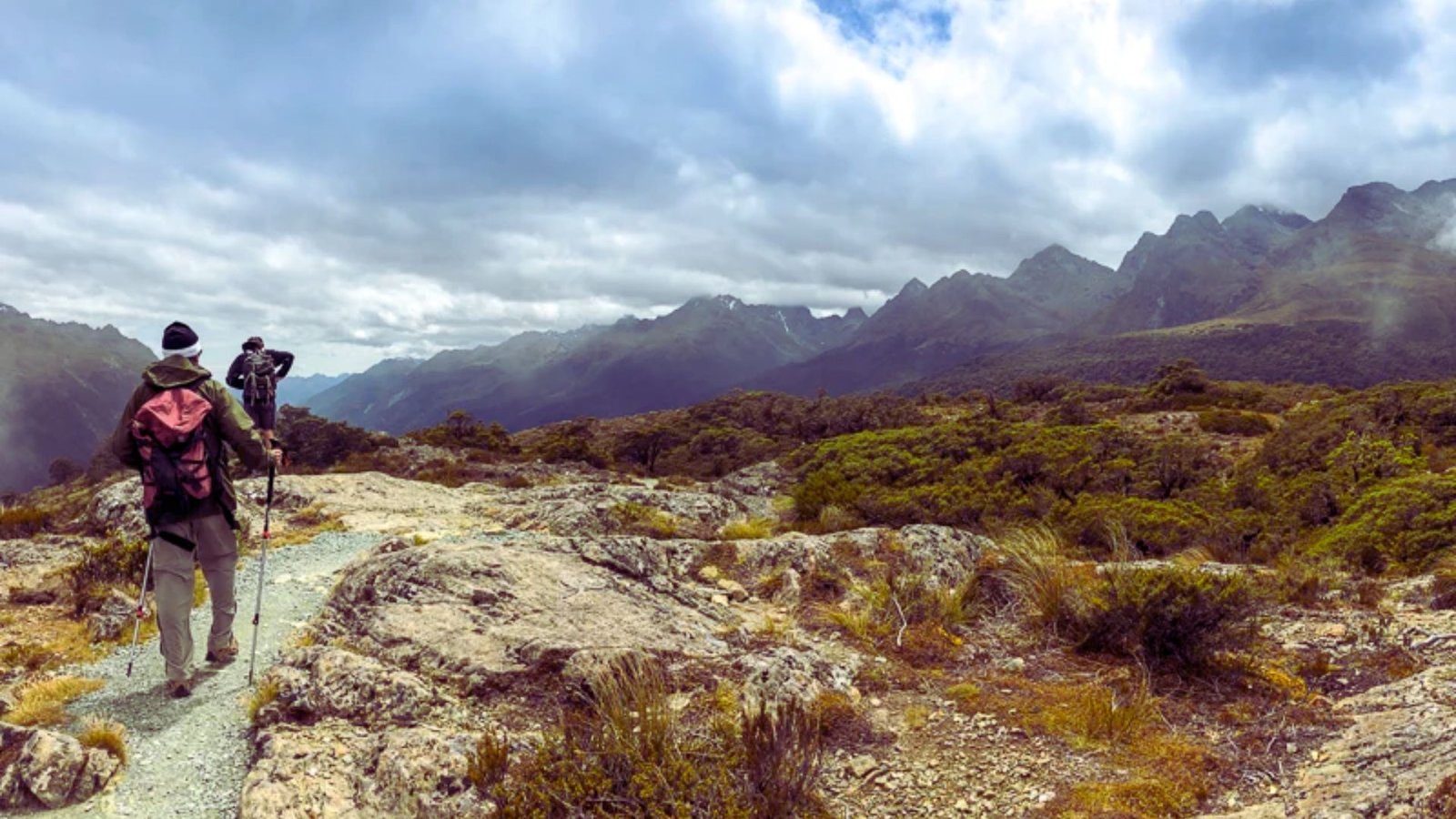 Two hikers with backpacks walking along a mountain trail surrounded by lush greenery and rocky terrain.